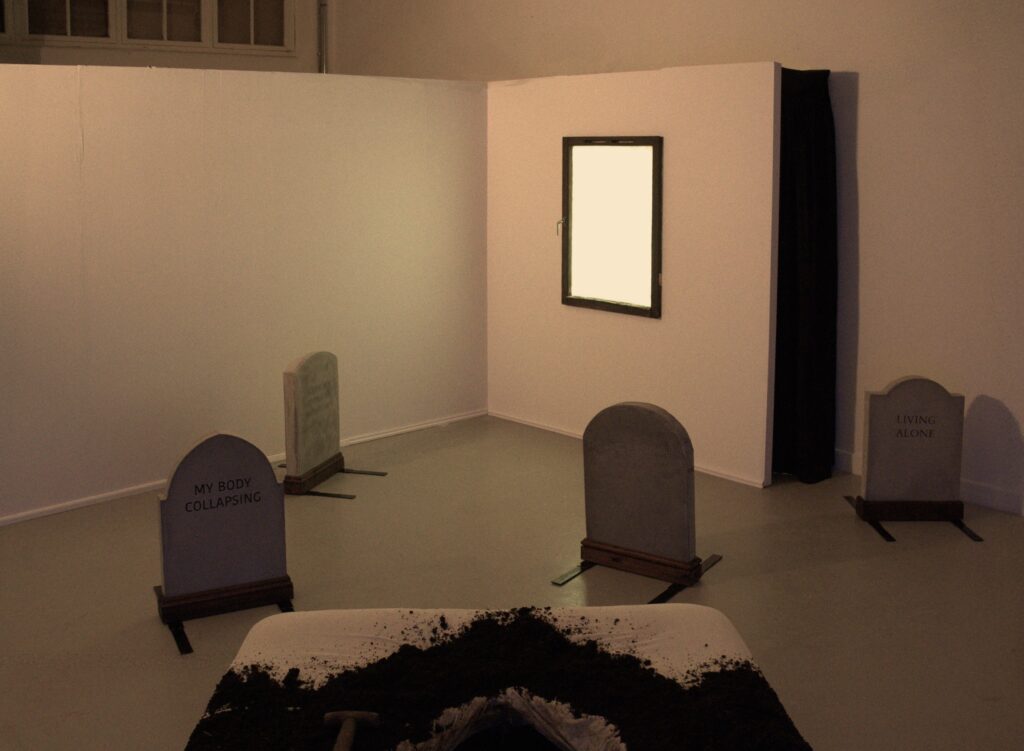 A bed with earth spilling out of it, facing four gravestones and an illuminated window.