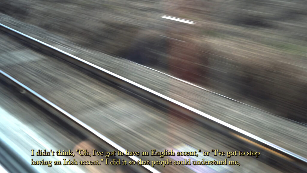 Blurred railway lines seen from a train window, overlaid with a subtitle about changing an Irish accent to be understood.