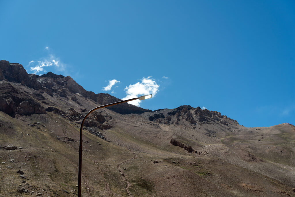 A streetlamp bends against the backdrop of a rocky mountain ridge under a clear blue sky.