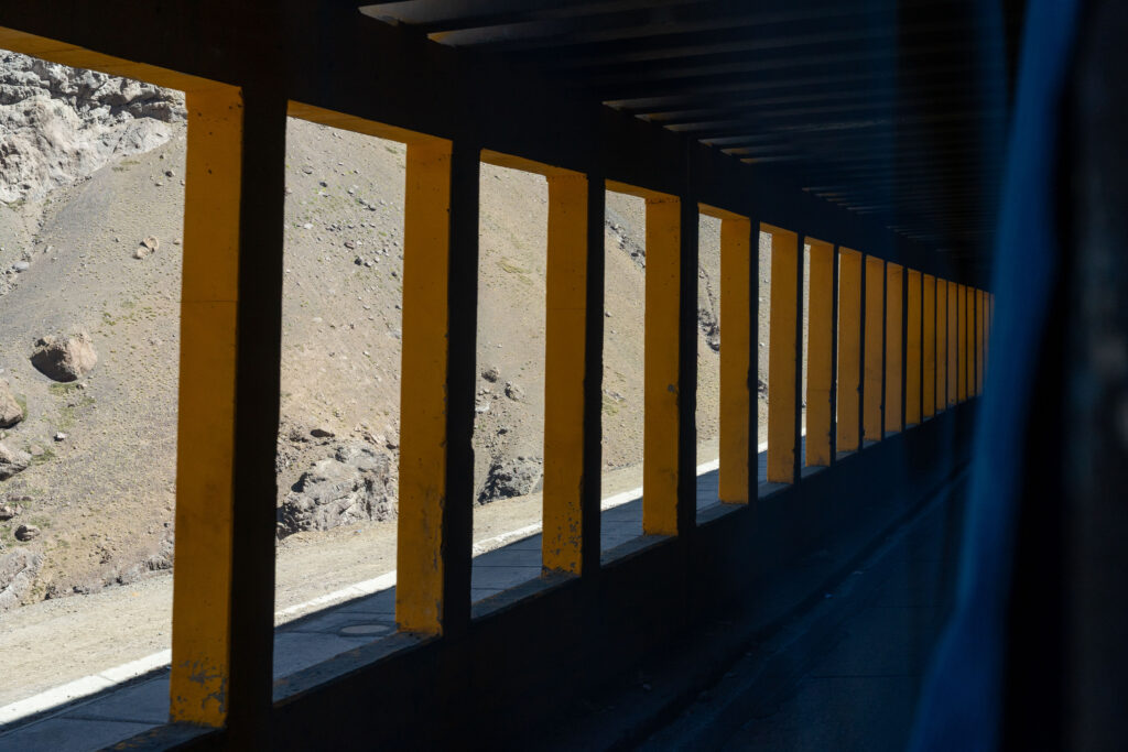 Yellow concrete supports form a repetitive colonnade beside a rocky hillside, seen from inside a bus.