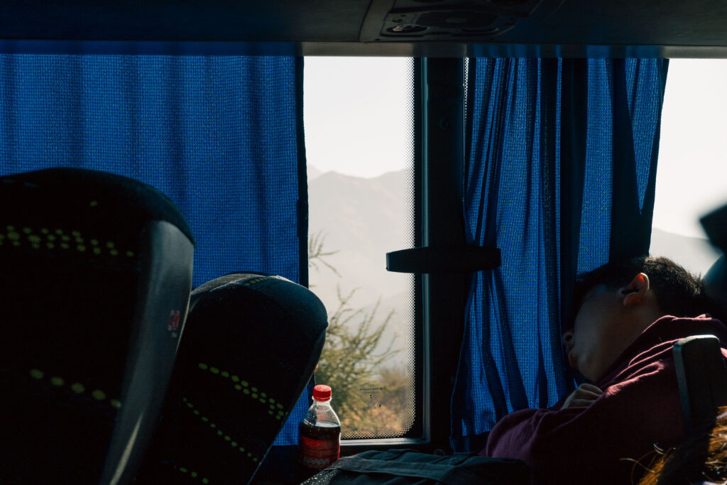 A young man sleeps against the window of a long-distance coach, blue curtain half drawn.